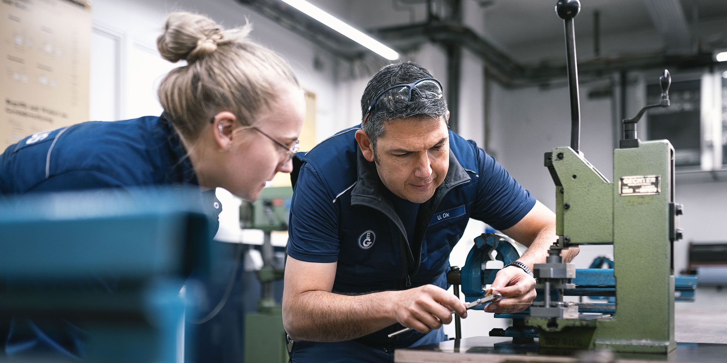 Ausbilder erklärt Auszubildender praktische Übung in Lehrwerkstatt, beide in Arbeitskleidung, praxisorientierte Ausbildung.